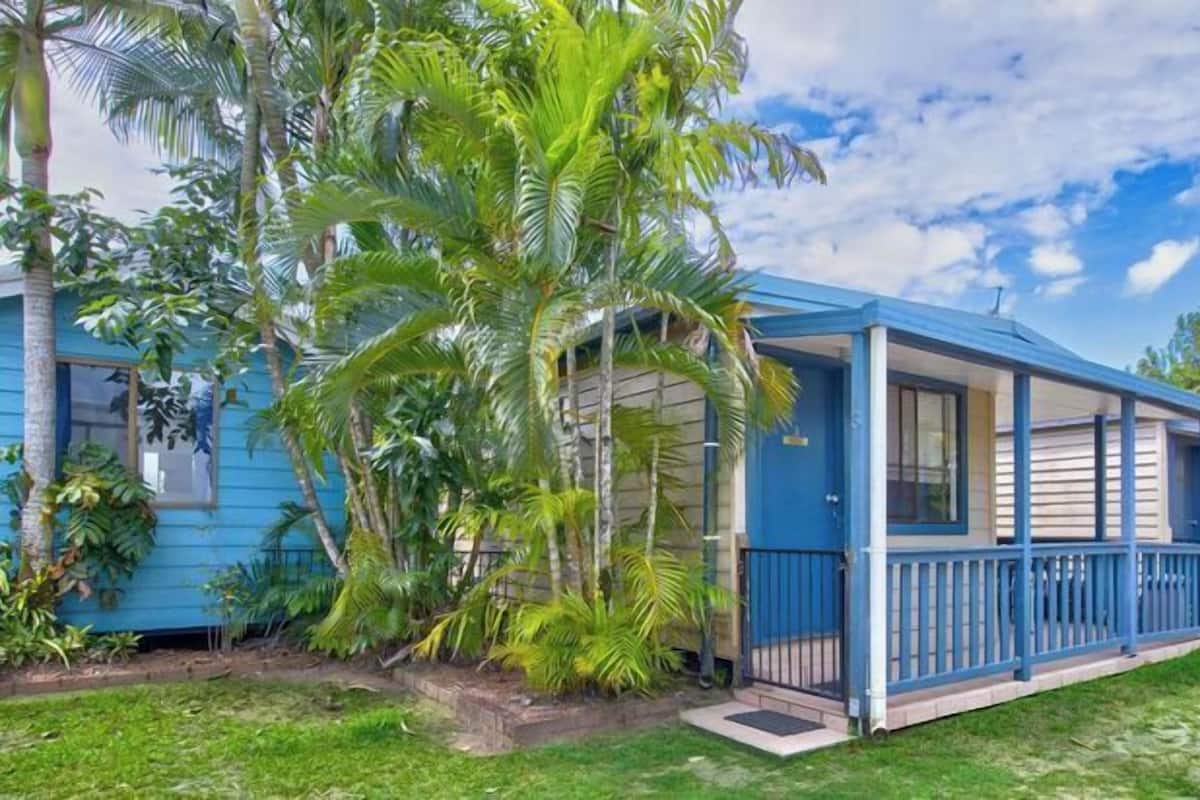 Una casa azul de dos pisos con porche, rodeada de vegetación tropical.