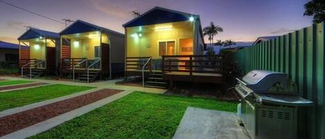 A row of small, single-story cabins with external stairs, a barbecue grill, and a fenced area.