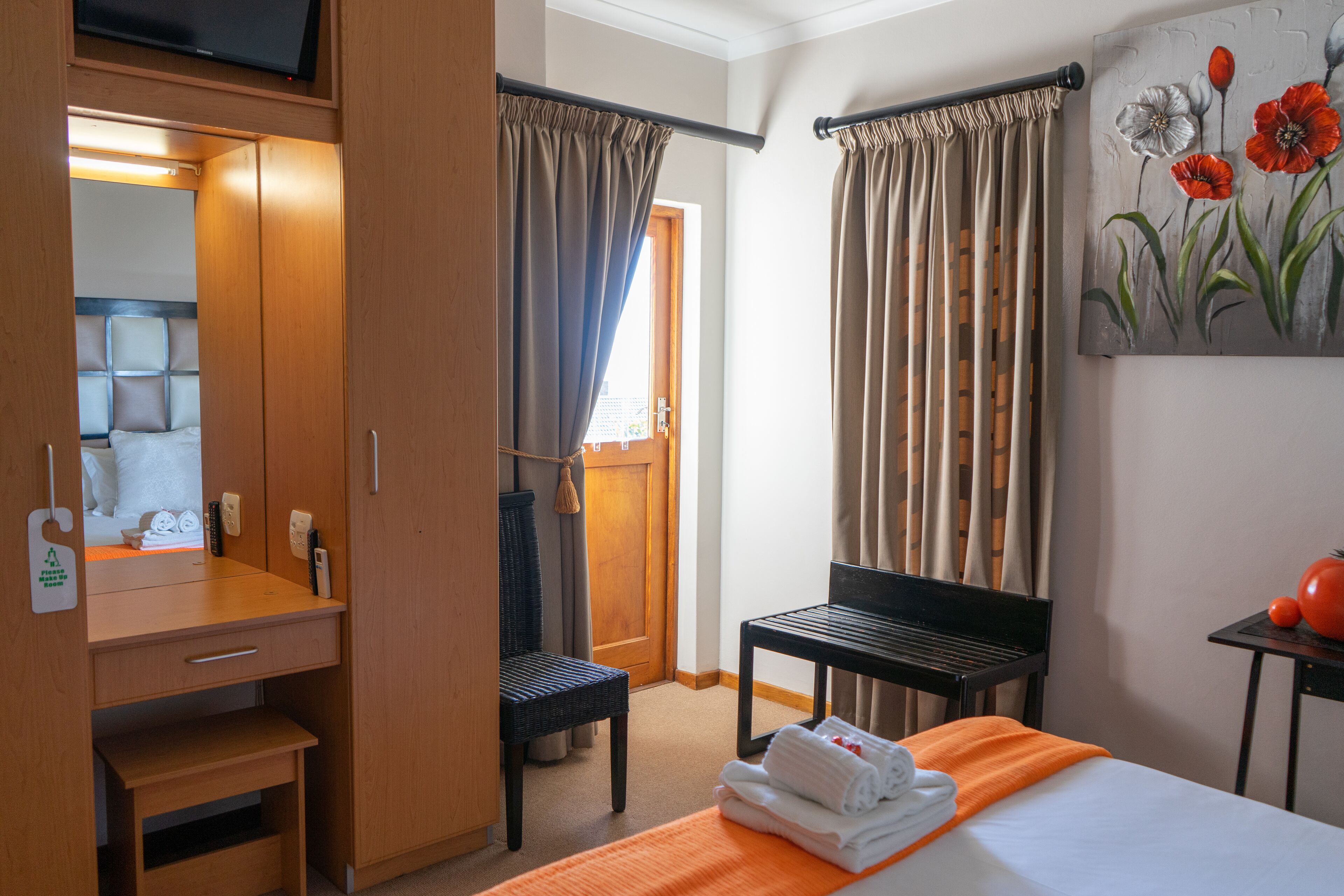 Habitación de hotel con un armario de madera, una cama con una colcha naranja, una silla, una mesita con un tomate y un cuadro de flores en la pared.