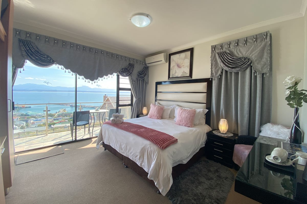 A hotel room with a large bed, a bedside table with a lamp, a view of the sea, and a balcony with chairs.