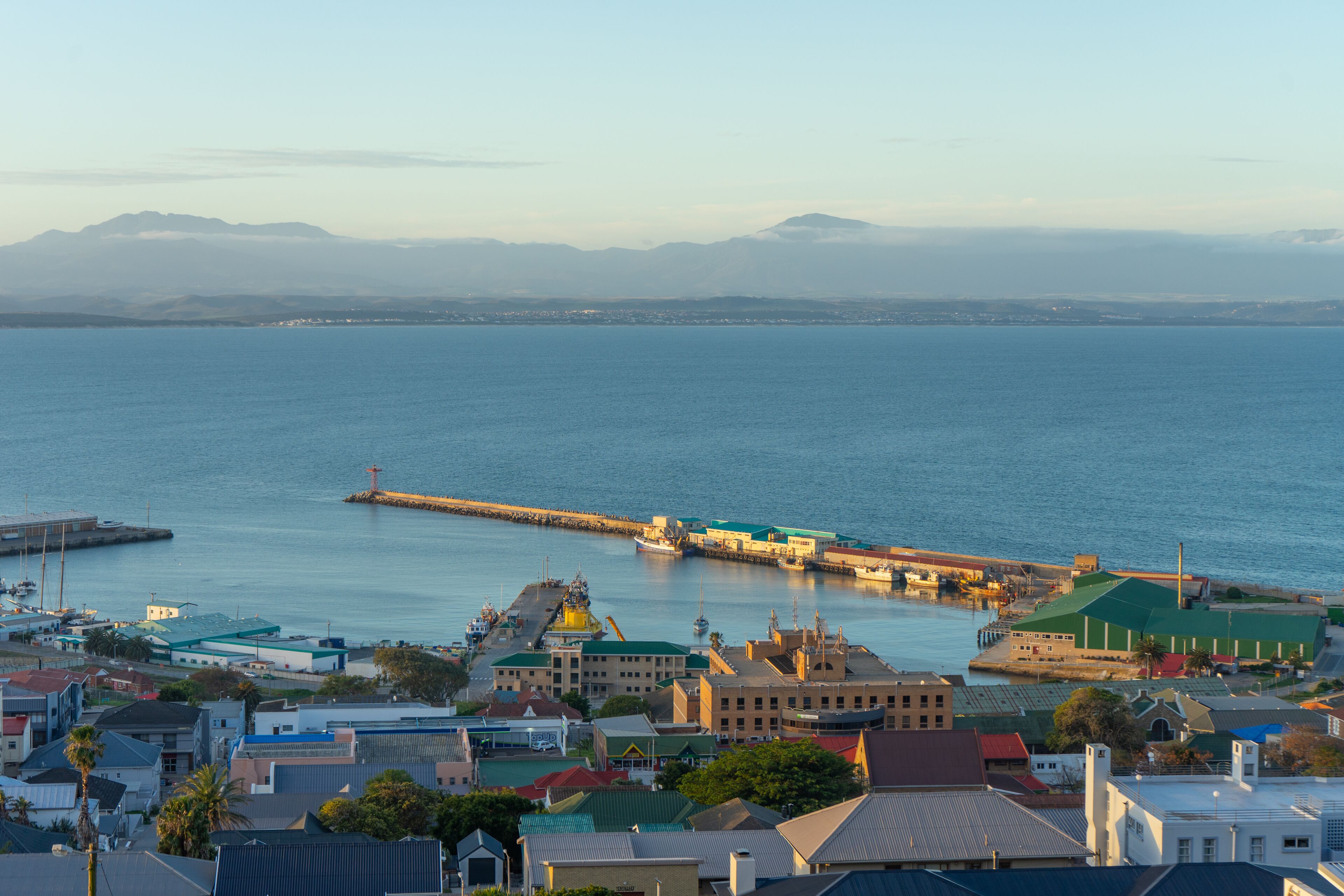 A coastal town with a prominent pier extending into the sea, surrounded by residential buildings and a clear view of distant mountains.