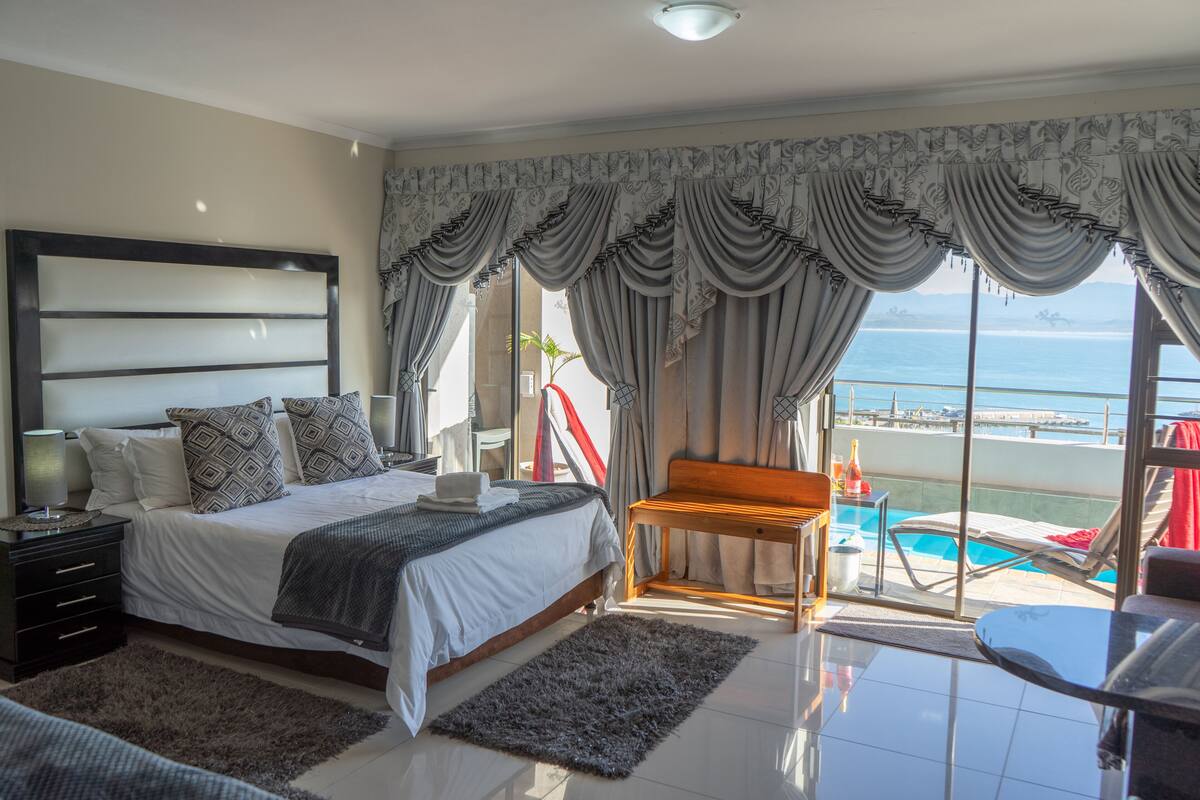 A bedroom with a large bed, a wooden bench, a small table, and a view of the sea through large windows.