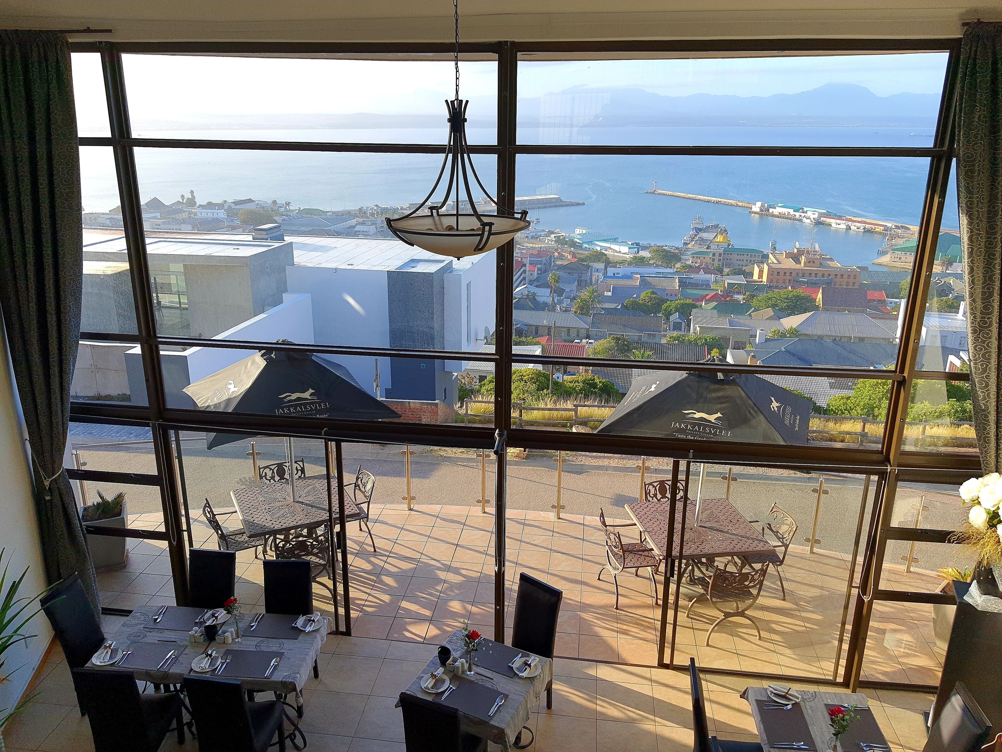 A dining area with tables and chairs, a hanging light fixture, and a view of a coastal cityscape through large windows.