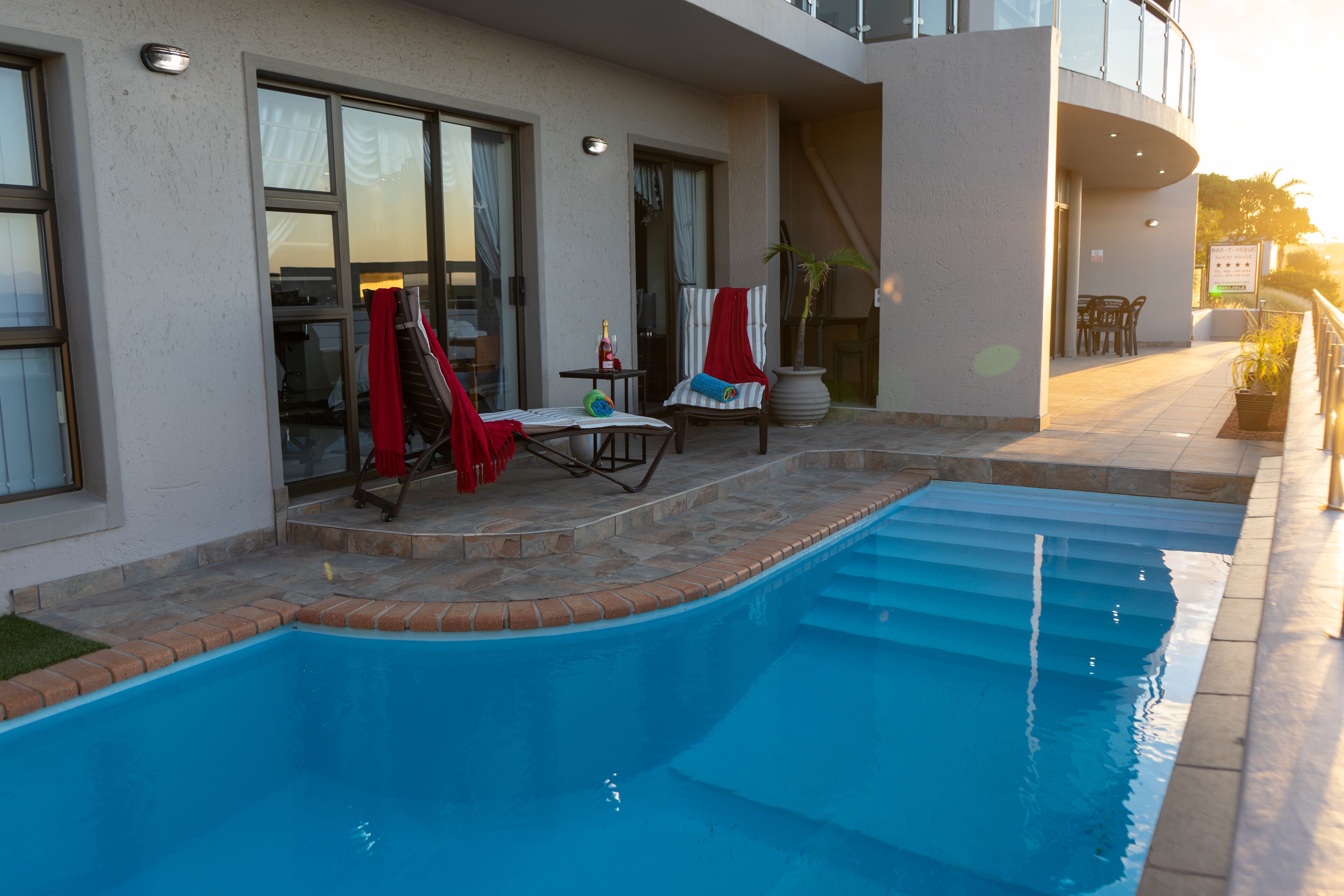 Una piscina con un patio soleggiato, una zona salotto coperta e una scala che conduce a un piano superiore.