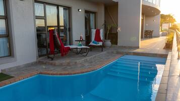 Una piscina con un patio soleggiato, una zona salotto coperta e una scala che conduce a un piano superiore.