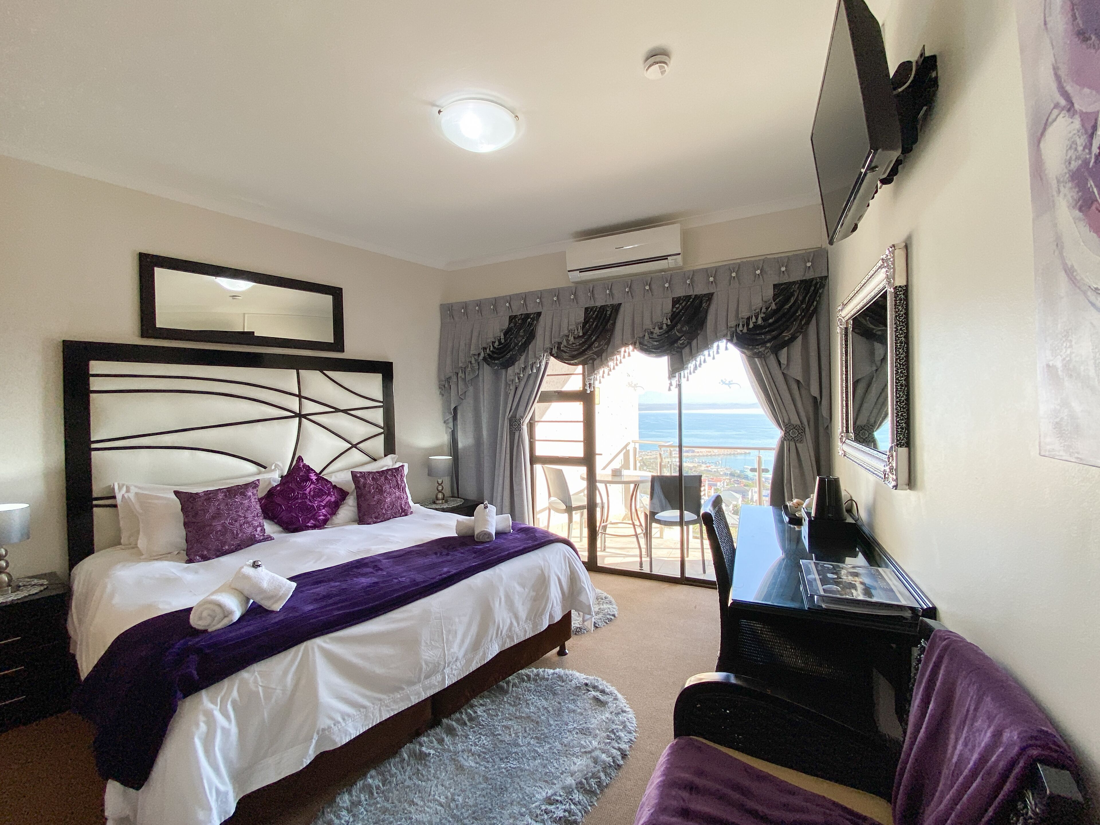 A hotel room with a bed, a desk, a chair, a TV, and a view of the sea.