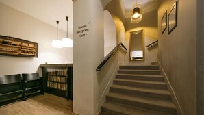 Zona de recepción con una escalera que conduce a los pisos superiores, un mostrador de recepción y un letrero en la pared que indica 'Recepción de habitaciones, Café'.