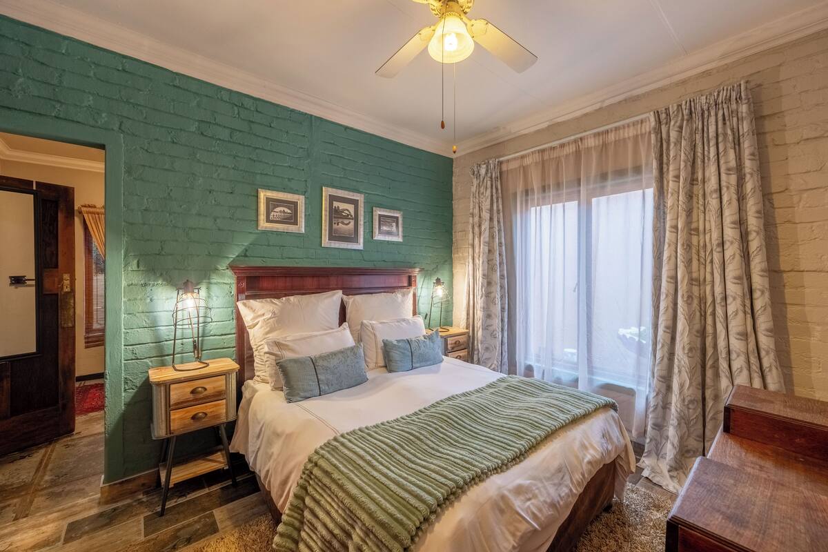 Quarto com parede de tijolos verdes, cabeceira de madeira, cama com roupa de cama branca, mesa de cabeceira com abajur e janela com cortinas leves.