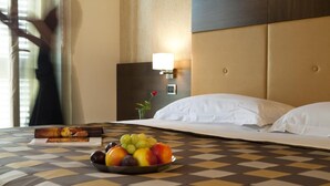 Een hotelkamer met een geruite sprei, een nachtkastje met een lamp, een boek en een schaal fruit.