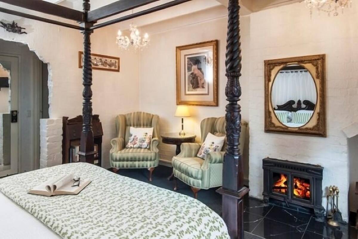 A four-poster bed, two armchairs, a fireplace, and a mirror in a room with a white brick wall and a chandelier.