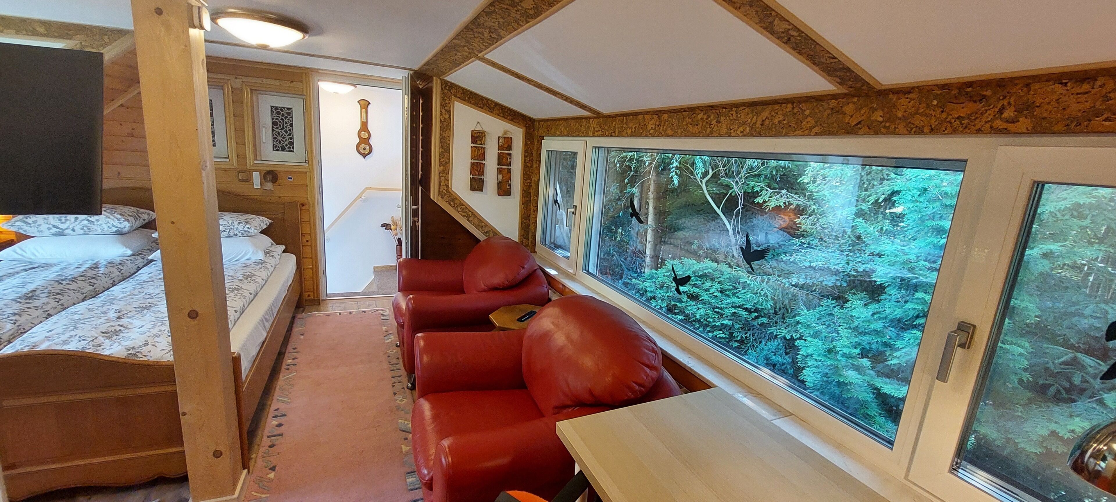 A cozy interior with a bed, a red leather couch, and a large window offering a forest view.