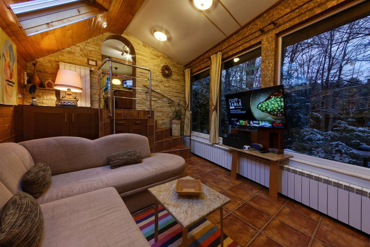 Ein gemütliches Wohnzimmer mit einer Couch, einem kleinen Tisch, einem Fernseher und einem Fenster mit Blick auf Bäume.