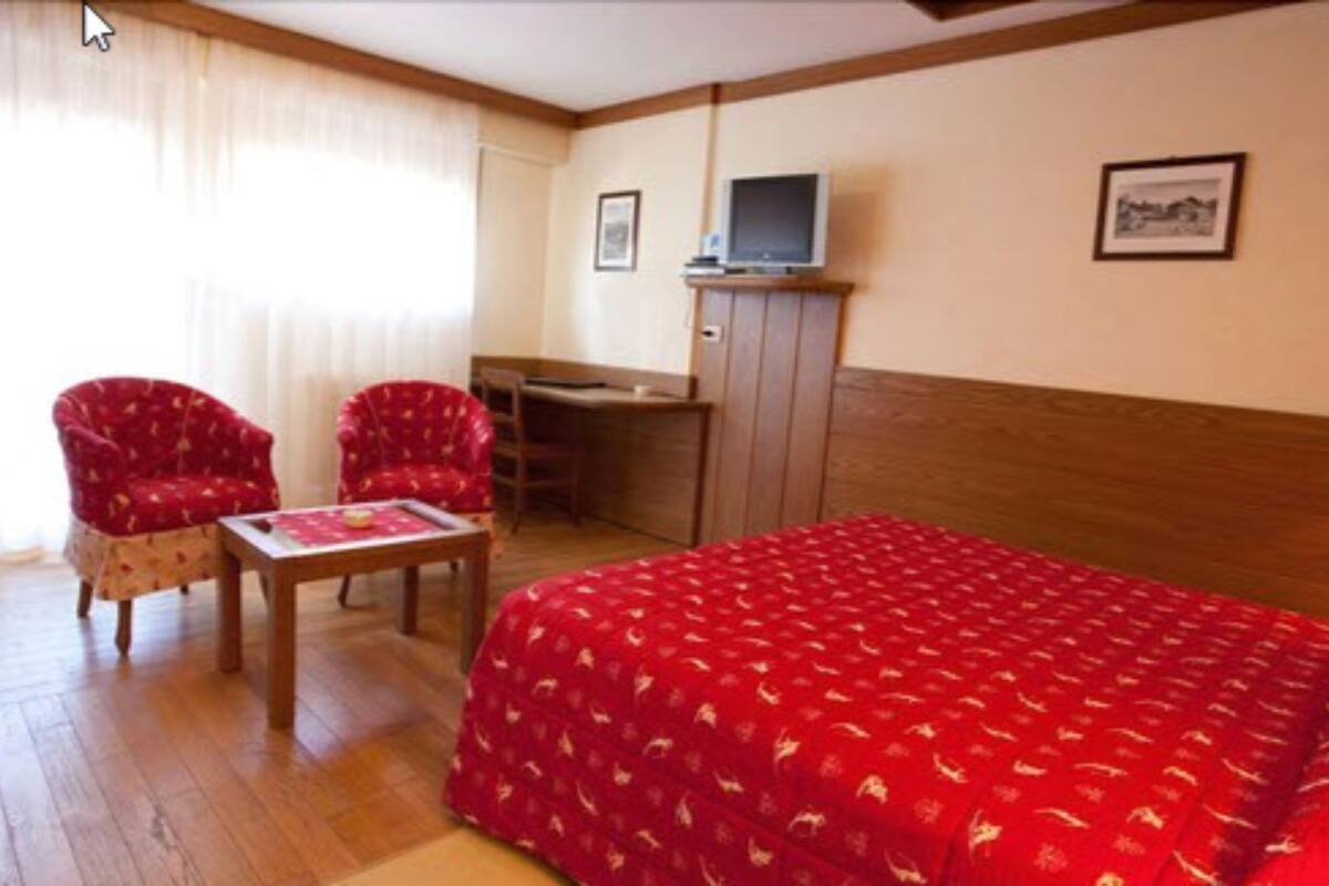 Habitación de hotel con una cama, dos sillones rojos, un escritorio de madera, un televisor y un cuadro en la pared.