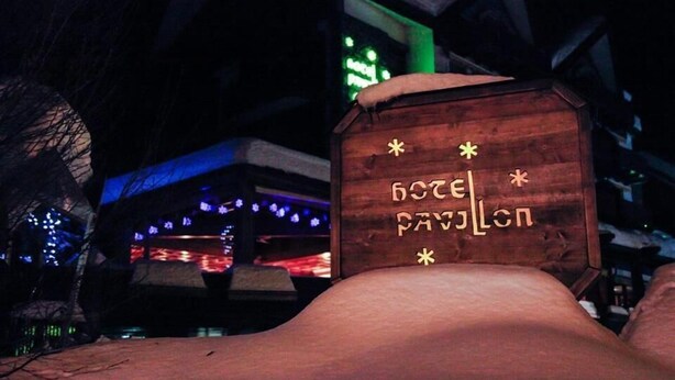 A wooden sign with the text "HOTEL PAVILION" and decorative stars, set against a snowy background with a building featuring blue lights in the background. - Hotel Pavillon (Courmayeur)