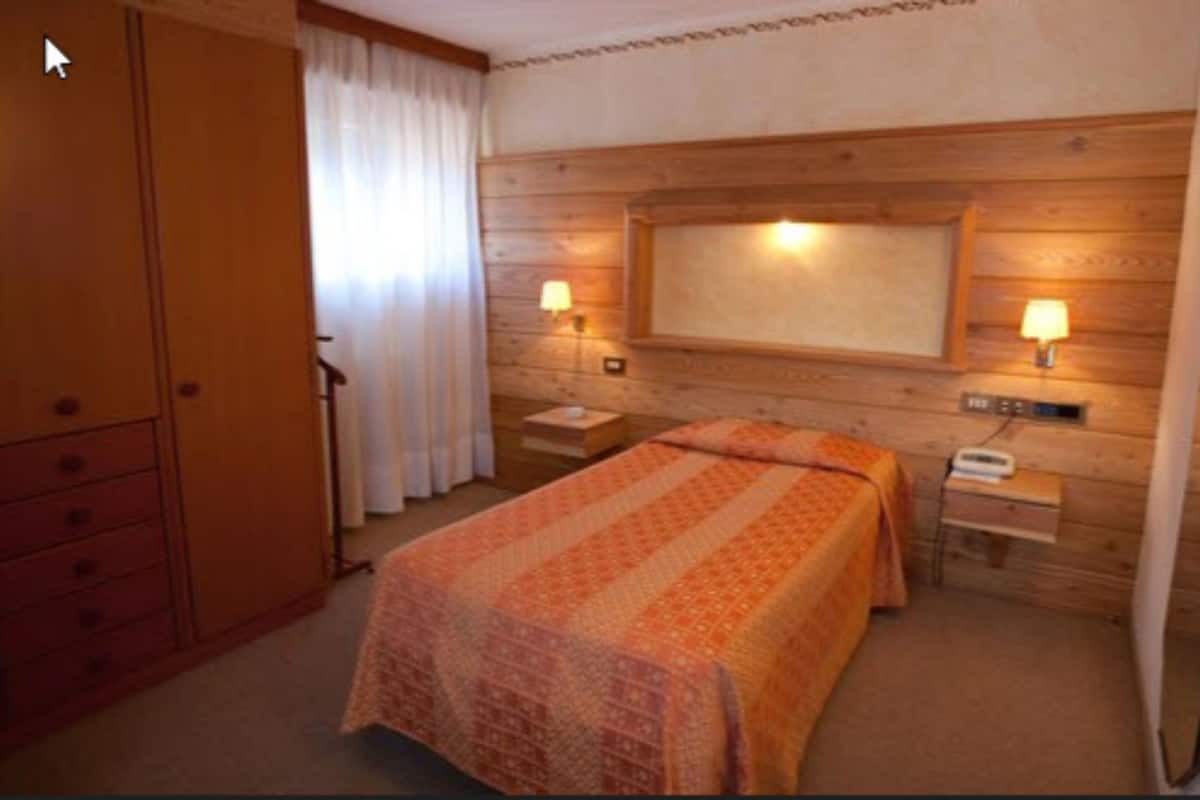 Habitación de hotel con pared de madera, una cama con colcha estampada, dos mesitas de noche con lámparas y un armario.
