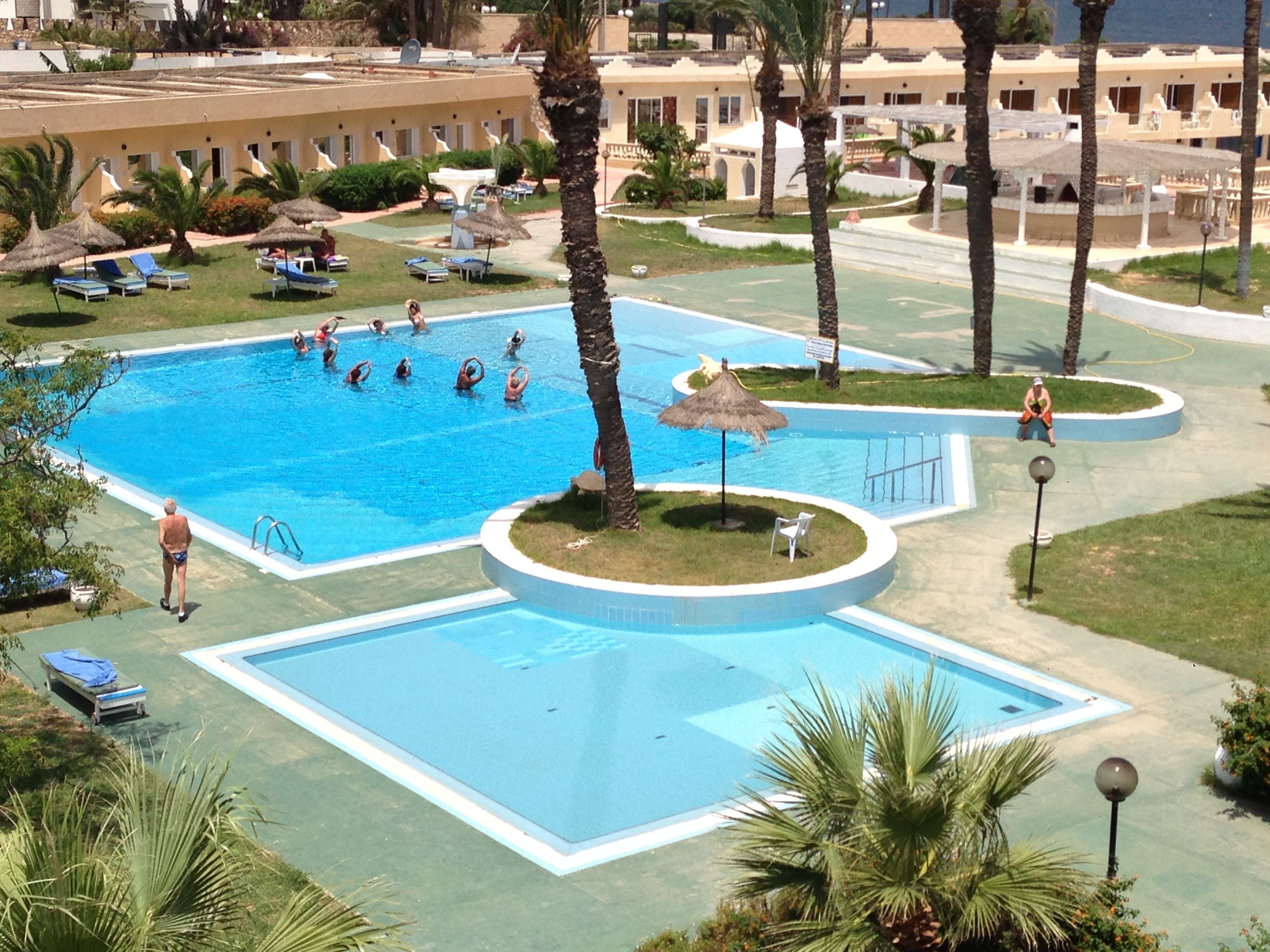 Una amplia piscina al aire libre rodeada de palmeras y sillas de descanso, con un edificio al fondo.
