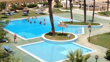 Una amplia piscina al aire libre rodeada de palmeras y sillas de descanso, con un edificio al fondo.