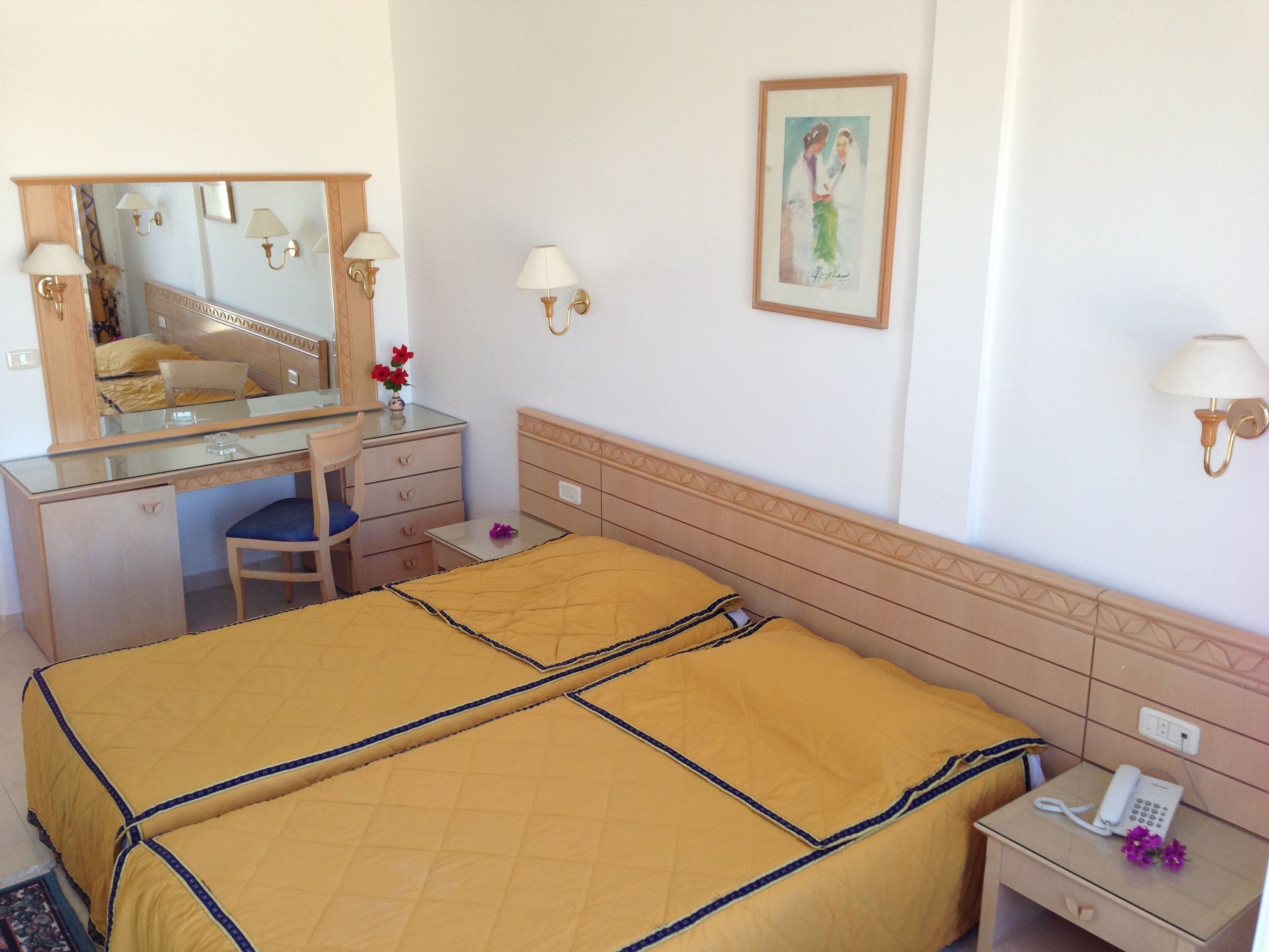 Habitación de hotel con cama, un escritorio con silla, un espejo, una mesita de noche con teléfono, y una lámpara fijada en la pared.
