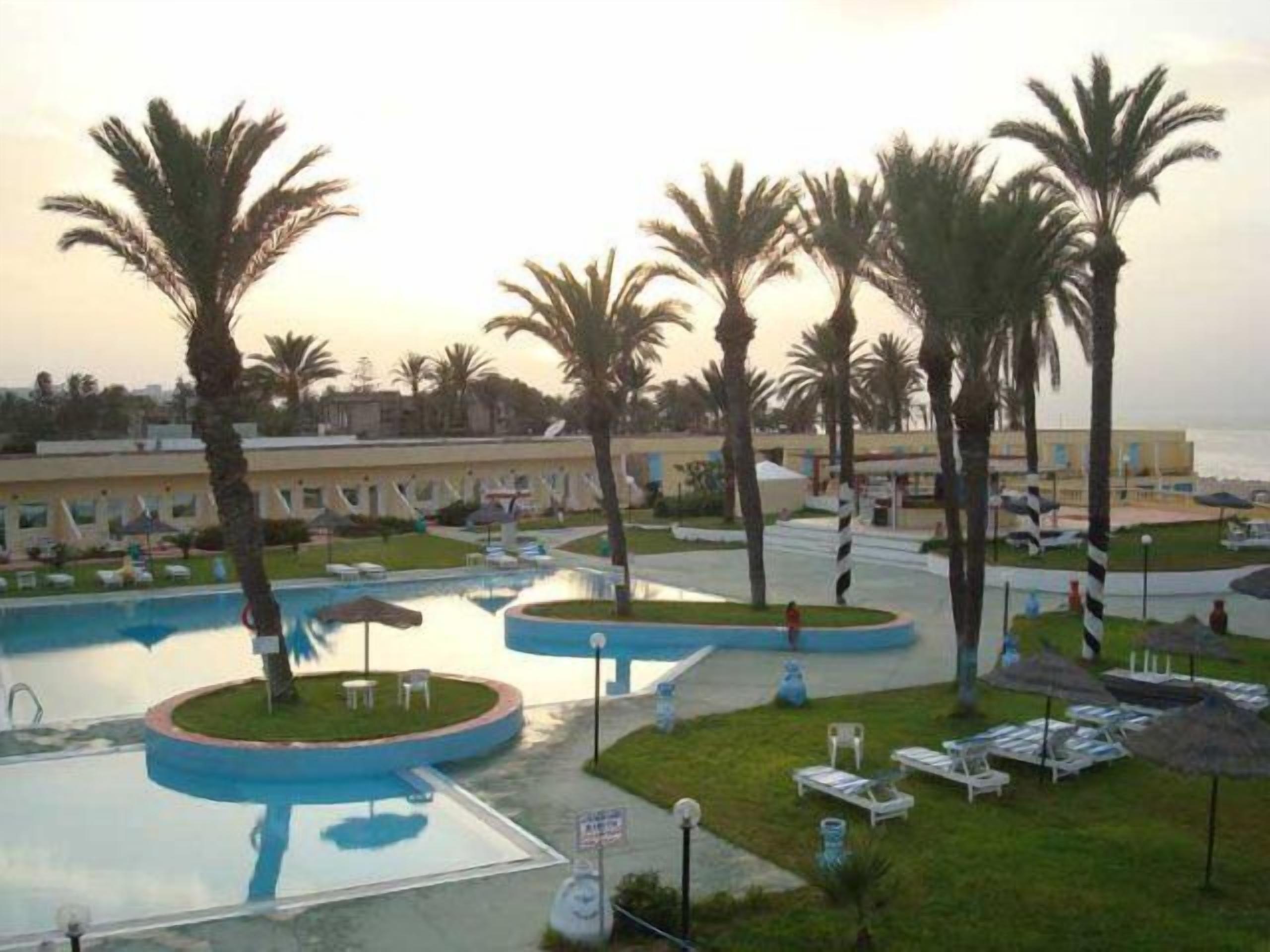 Ein Poolbereich eines Resorts mit Palmen, Liegestühlen und einem Gebäude im Hintergrund.