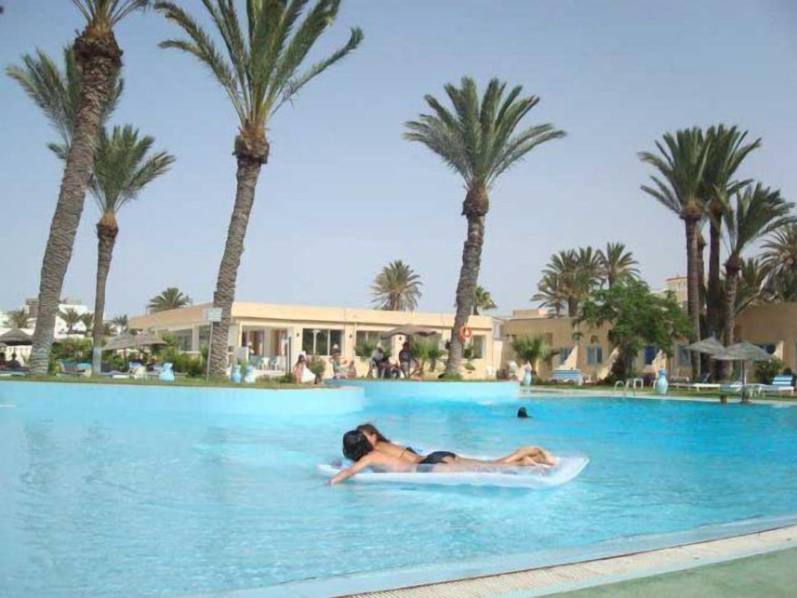 A pool area with palm trees, lounge chairs, and a building in the background.