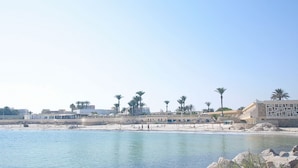 Una zona costera con playa de arena, palmeras y un edificio de fachada geométrica.