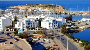 Un pueblo costero con edificios blancos, un puerto deportivo con barcos y una colina rocosa al fondo.