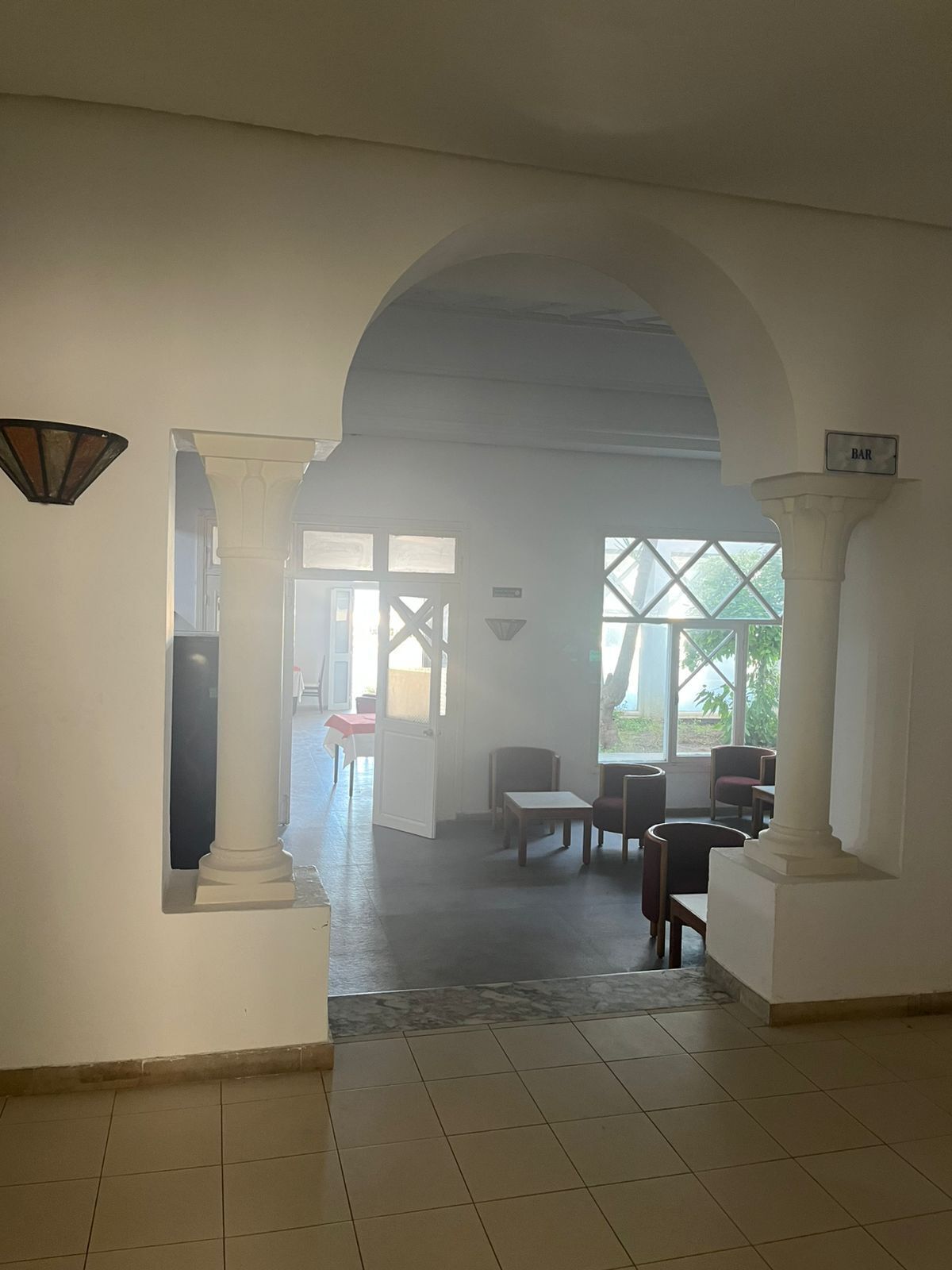 Un couloir avec des colonnes blanches, une enseigne de bar et une vue sur un espace extérieur aménagé avec des tables et des chaises.