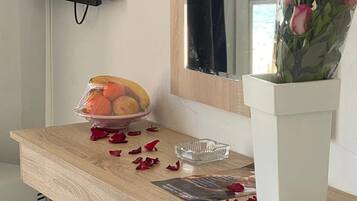Et træskrivebord med en skål med frugt, en glasaskebæger og en vase med blomster. Et fjernsyn er monteret på væggen over skrivebordet.
