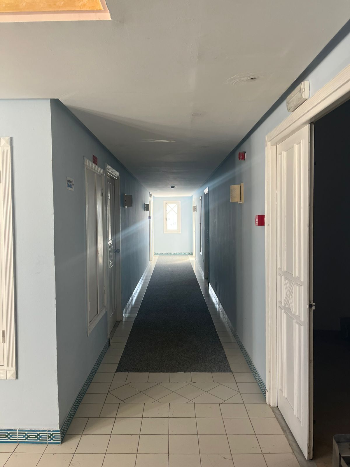 Un long couloir étroit aux murs bleu clair, aux portes blanches et au tapis foncé.
