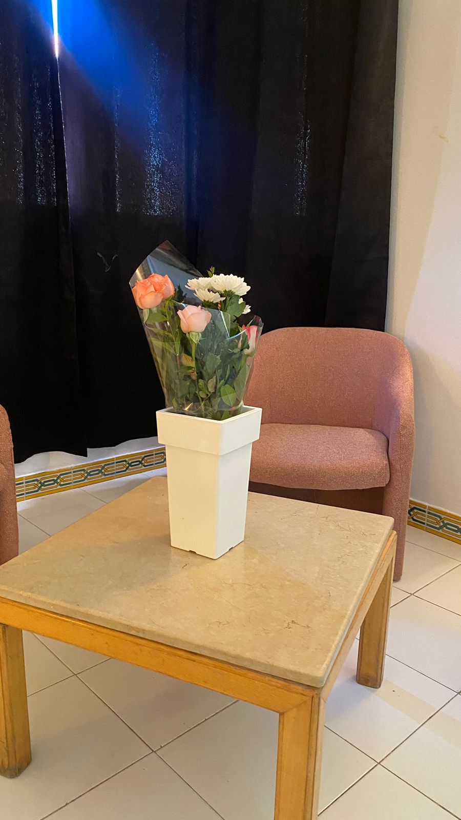 Eine weiße Vase mit Blumen auf einem kleinen Tisch in einem Raum mit schwarzem Vorhang im Hintergrund.