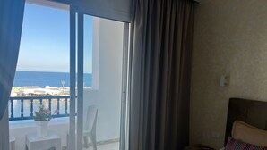 Una habitación de hotel con cama, mesita de noche, ventana con vistas al mar y balcón con una silla y una pequeña mesa.