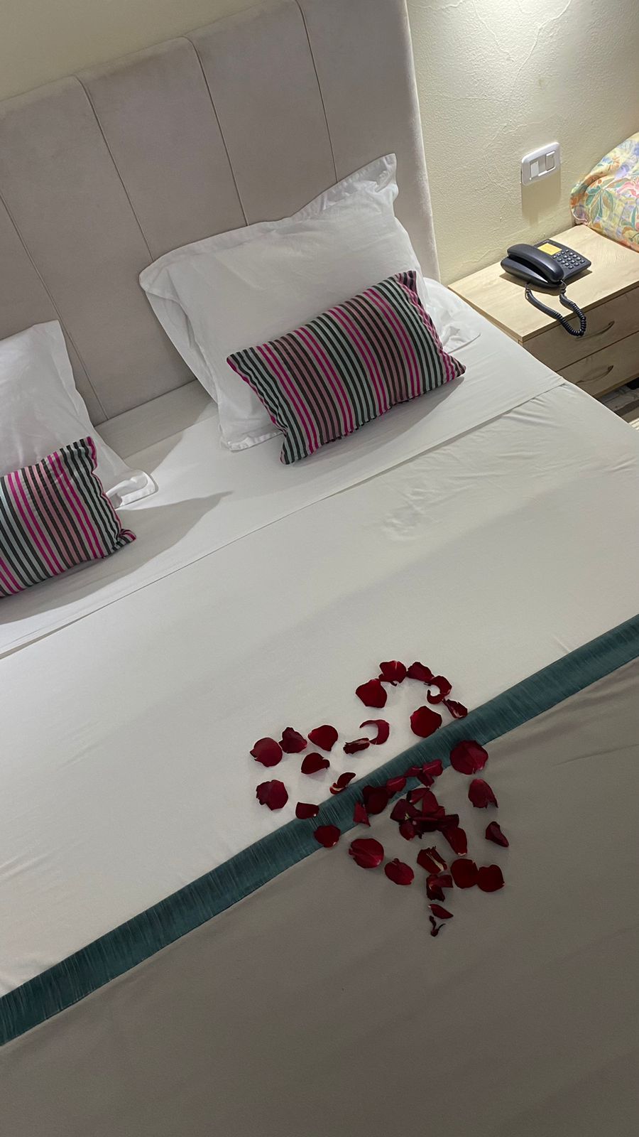 Et hotelværelse med en seng, to puder, et natbord med en telefon og rosenblade på sengen.