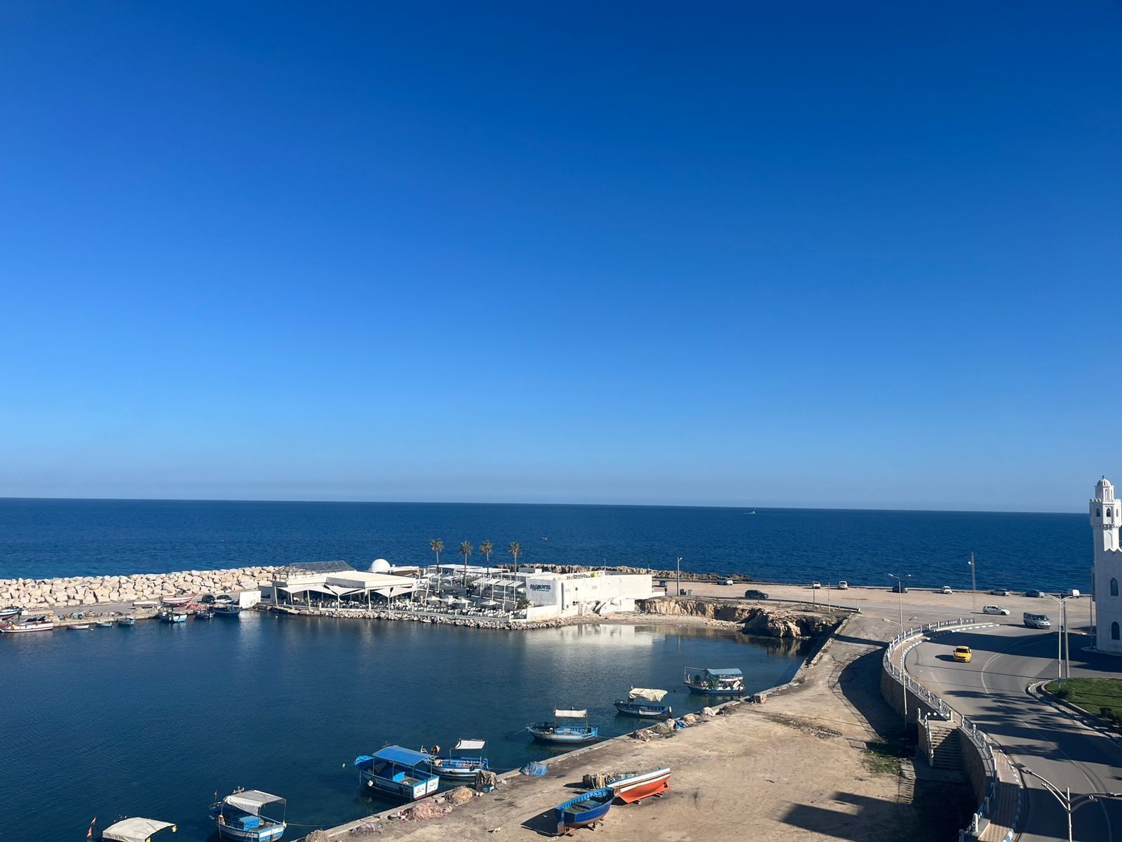 A coastal marina with boats, a white building, and a road leading to a white tower.