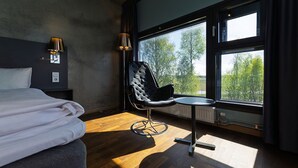 Une chambre d’hôtel moderne avec un grand lit, une chaise, une petite table et une vue sur les arbres à travers de grandes fenêtres.