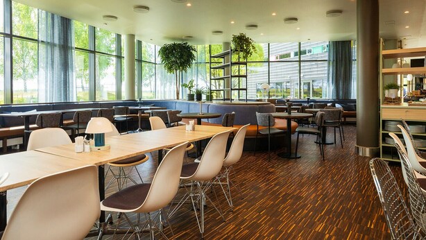 Un restaurant moderne doté de grandes fenêtres, d’un plancher en bois et proposant différents types de sièges, notamment des tables et des banquettes.