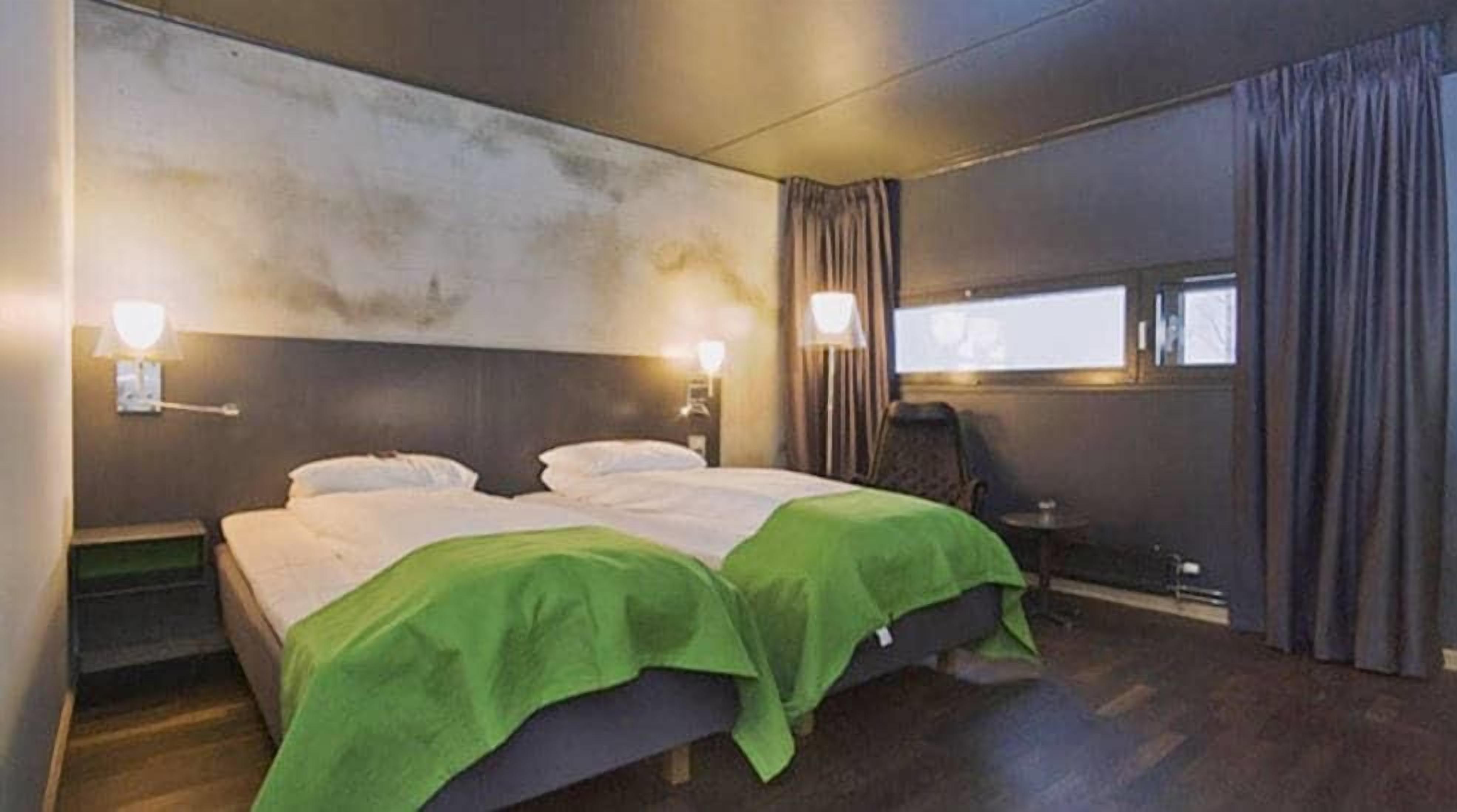 A hotel room with a bed, two bedside lamps, a window with curtains, and a small table.