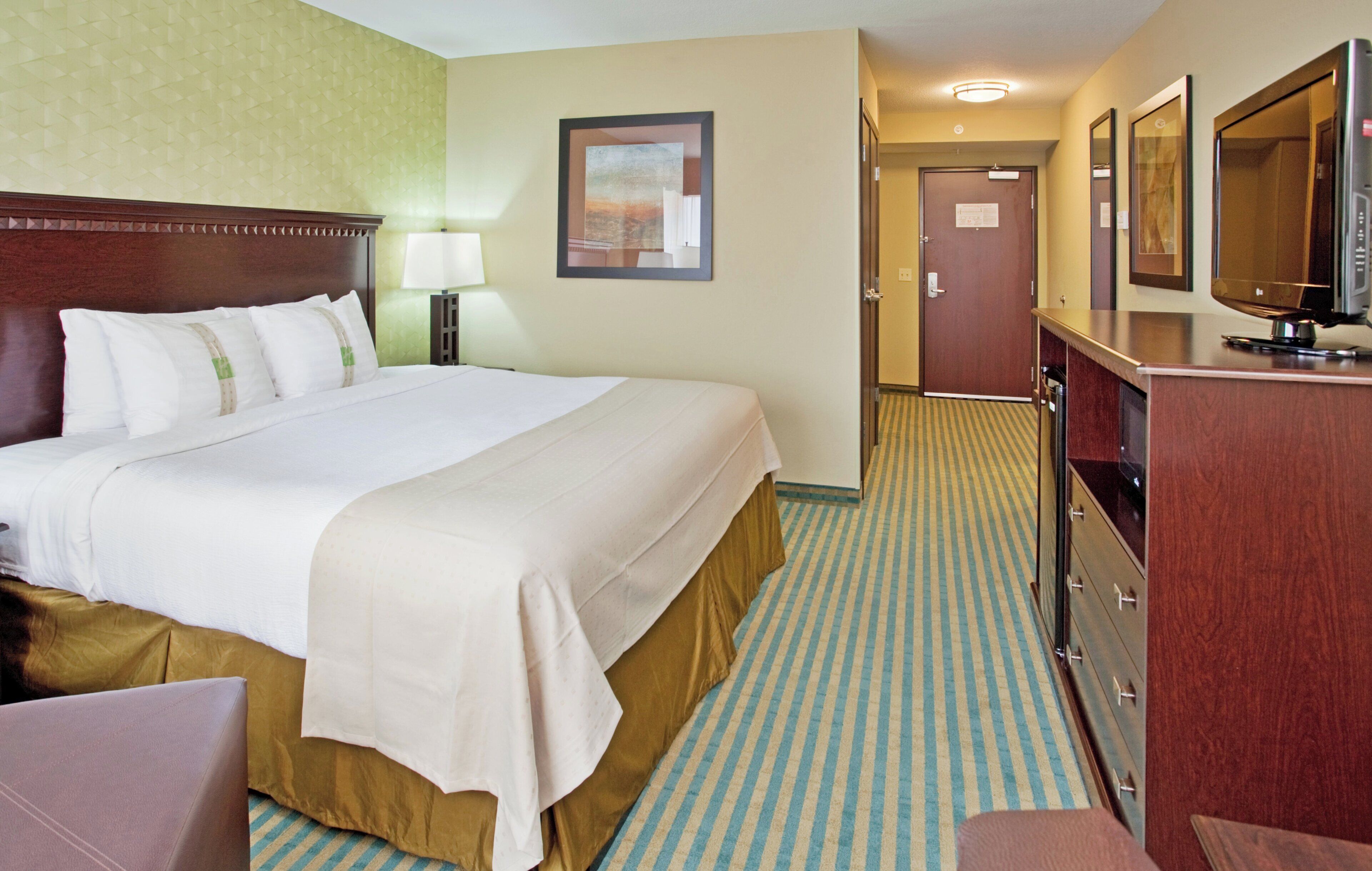 Une chambre d’hôtel avec un grand lit, un bureau doté d’un téléviseur à écran plat, un miroir et une porte donnant sur un couloir.