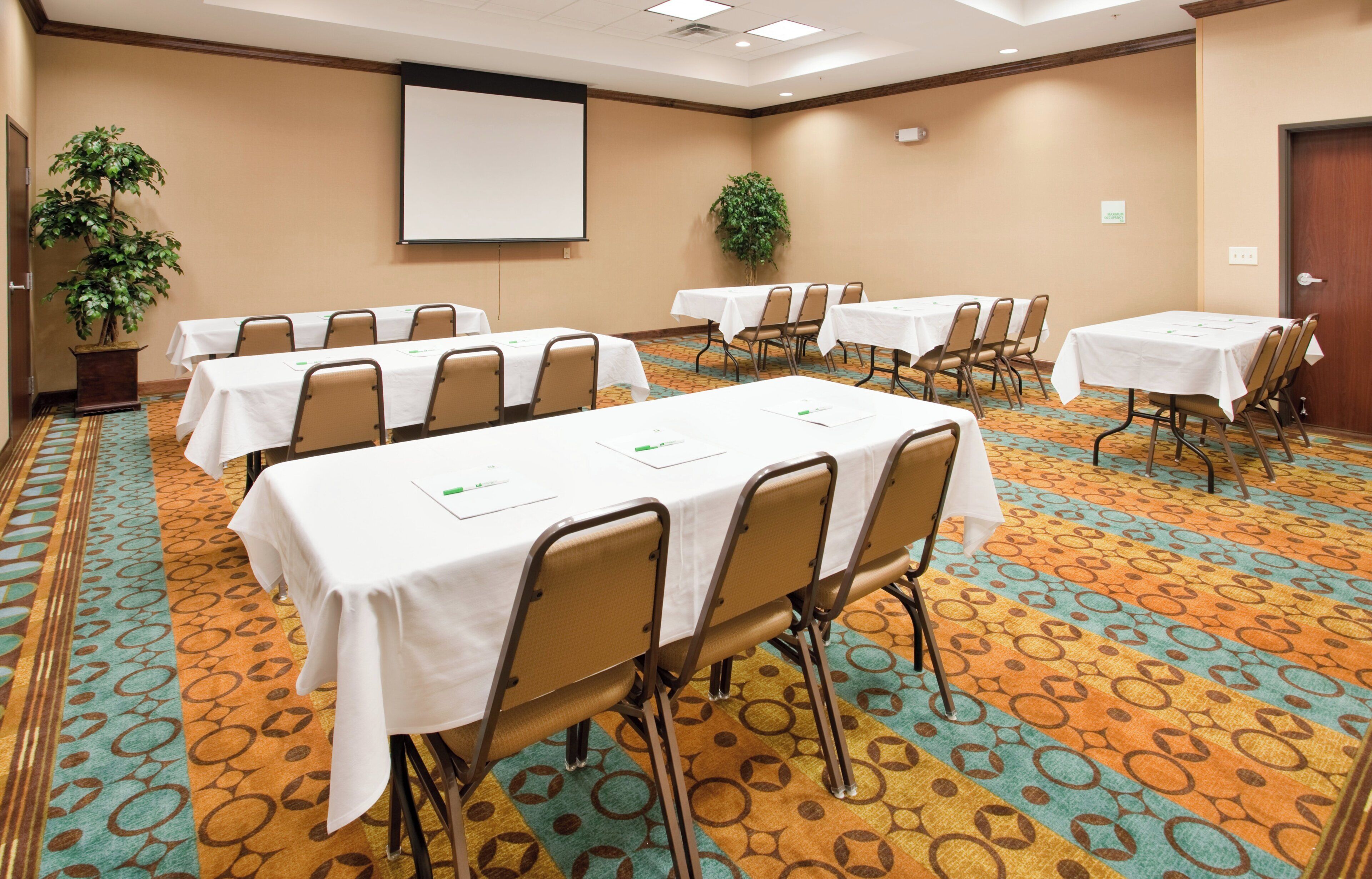 A conference room with multiple tables and chairs, a projector screen, and potted plants.