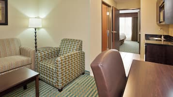Hotelværelse med sofa, lænestol, lille bord, lampe, tekøkken og seng med gardiner.