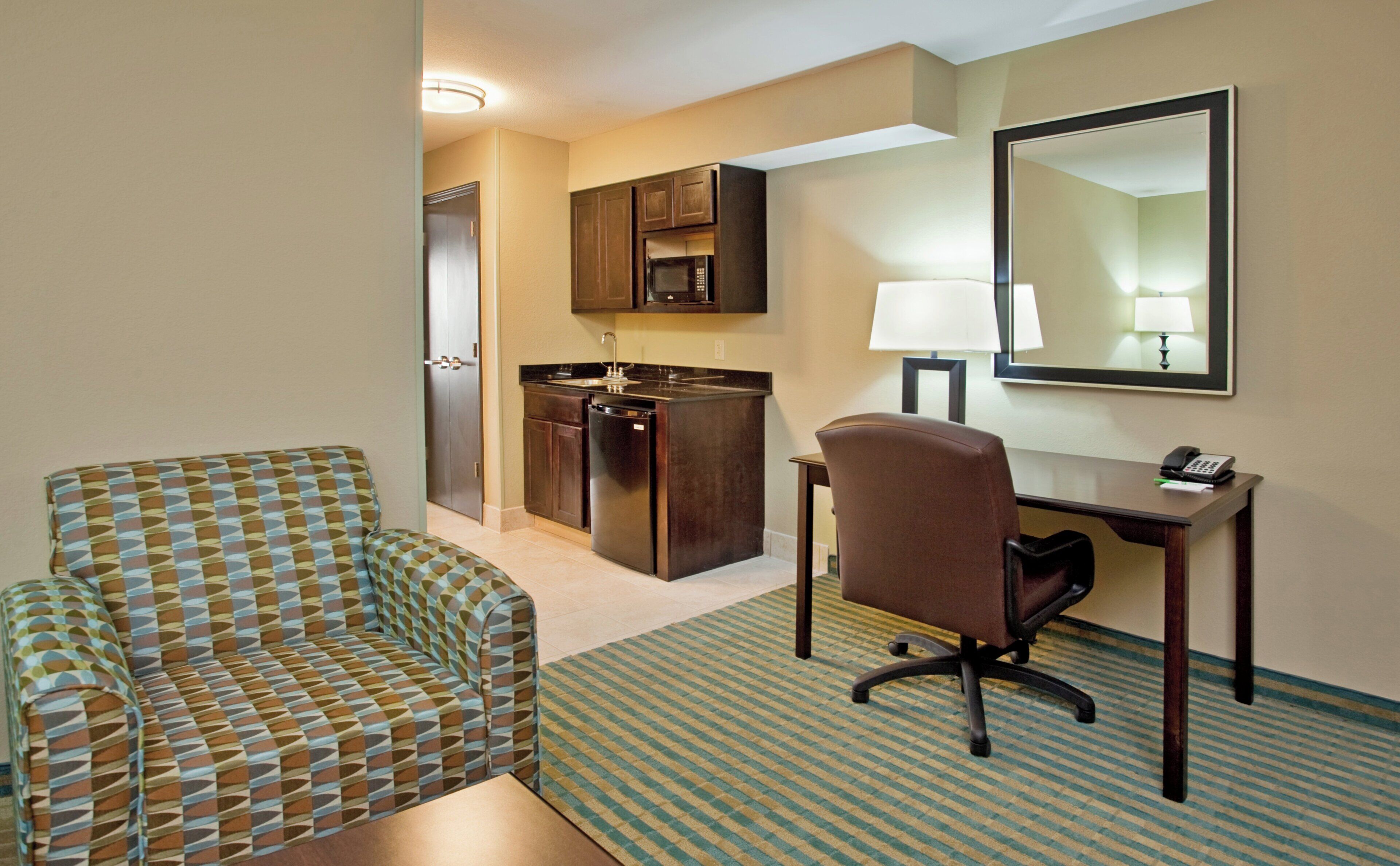 A hotel room with a patterned sofa, a desk with a lamp, a telephone, and a kitchen area with cabinets and a refrigerator.