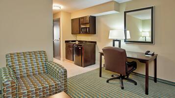 A hotel room with a patterned sofa, a desk with a lamp, a telephone, and a kitchen area with cabinets and a refrigerator.