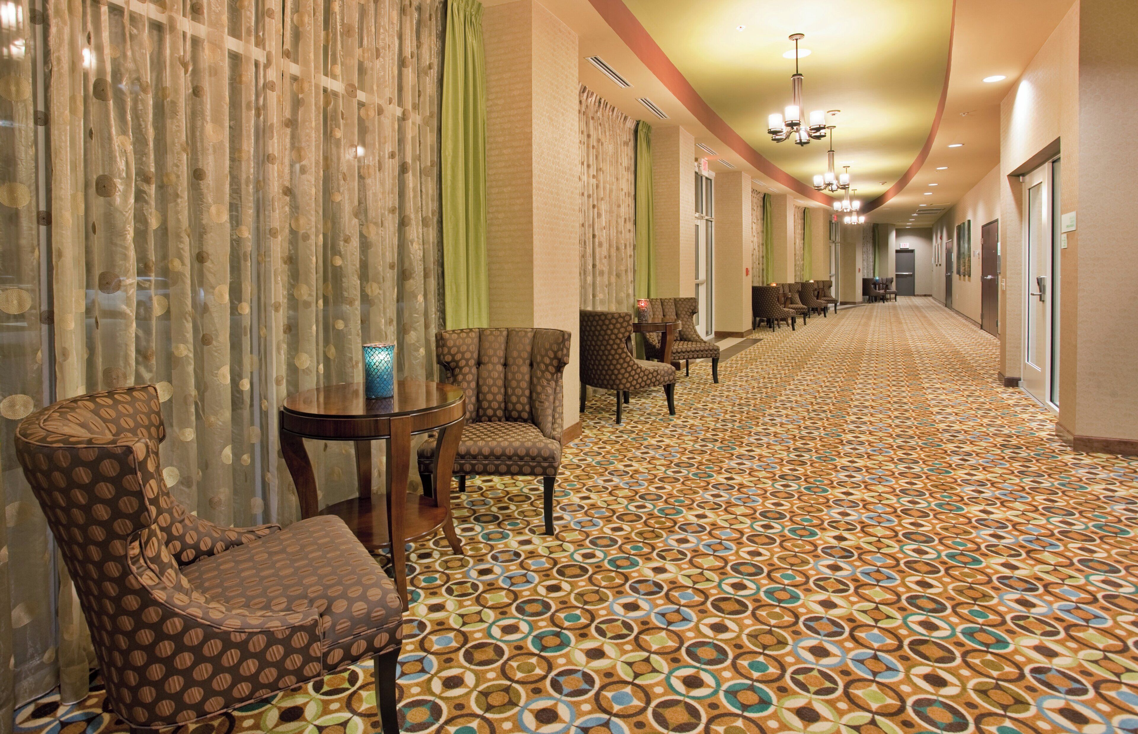 A hotel corridor with patterned carpet, seating area with chairs and a table, and a chandelier.