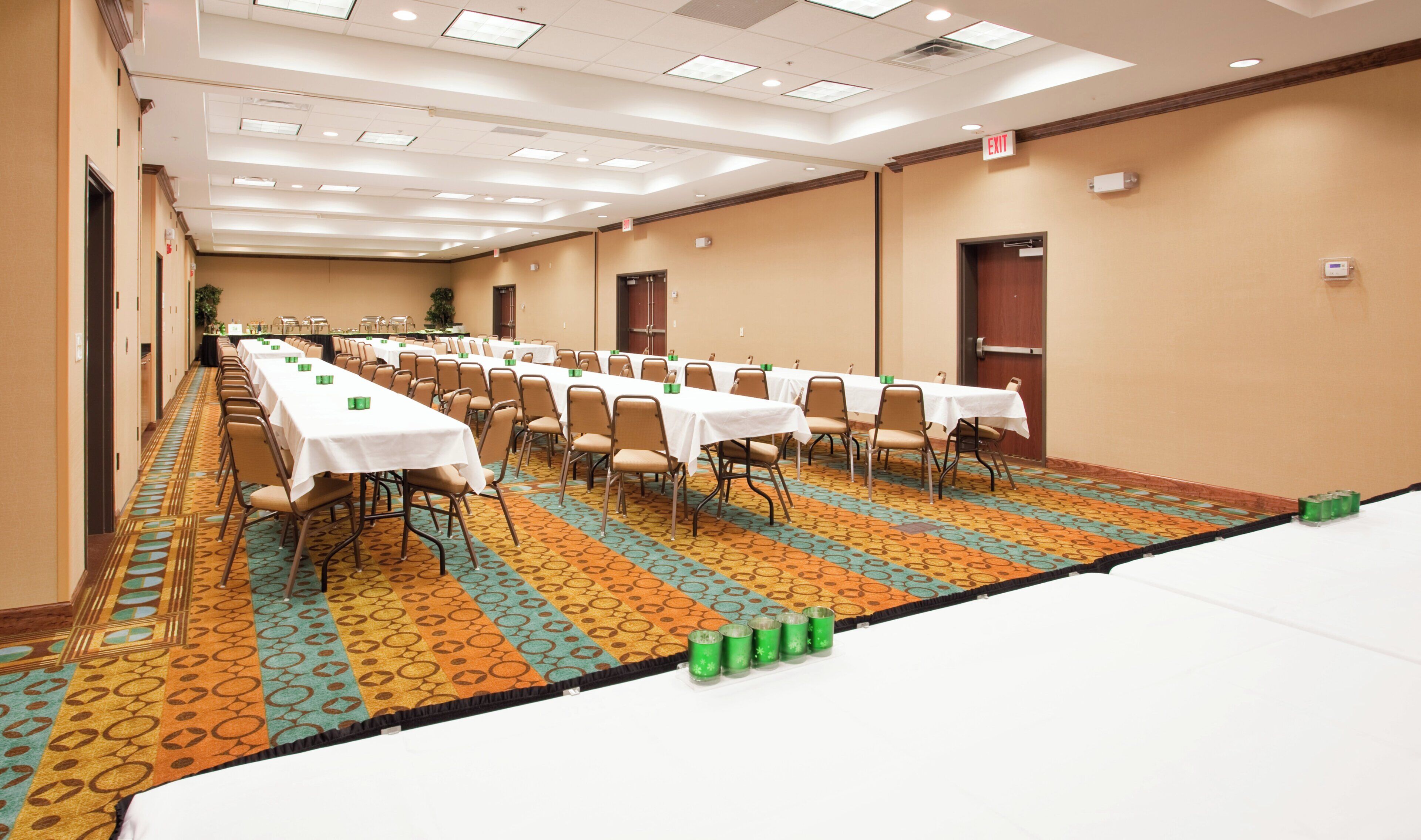 Un salón de conferencias con filas de mesas y sillas, piso alfombrado y señal de salida.