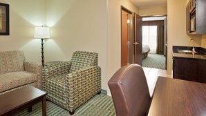 Ein Hotelzimmer mit einer Couch, einem Sessel und einem kleinen Tisch. Es gibt eine Lampe, ein Bett sowie eine Küchenzeile mit Spüle und Mikrowelle.