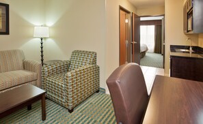 Une chambre d’hôtel avec un canapé, un fauteuil et une petite table. On y trouve une lampe, un lit et une kitchenette équipée d’un évier et d’un four à micro-ondes.