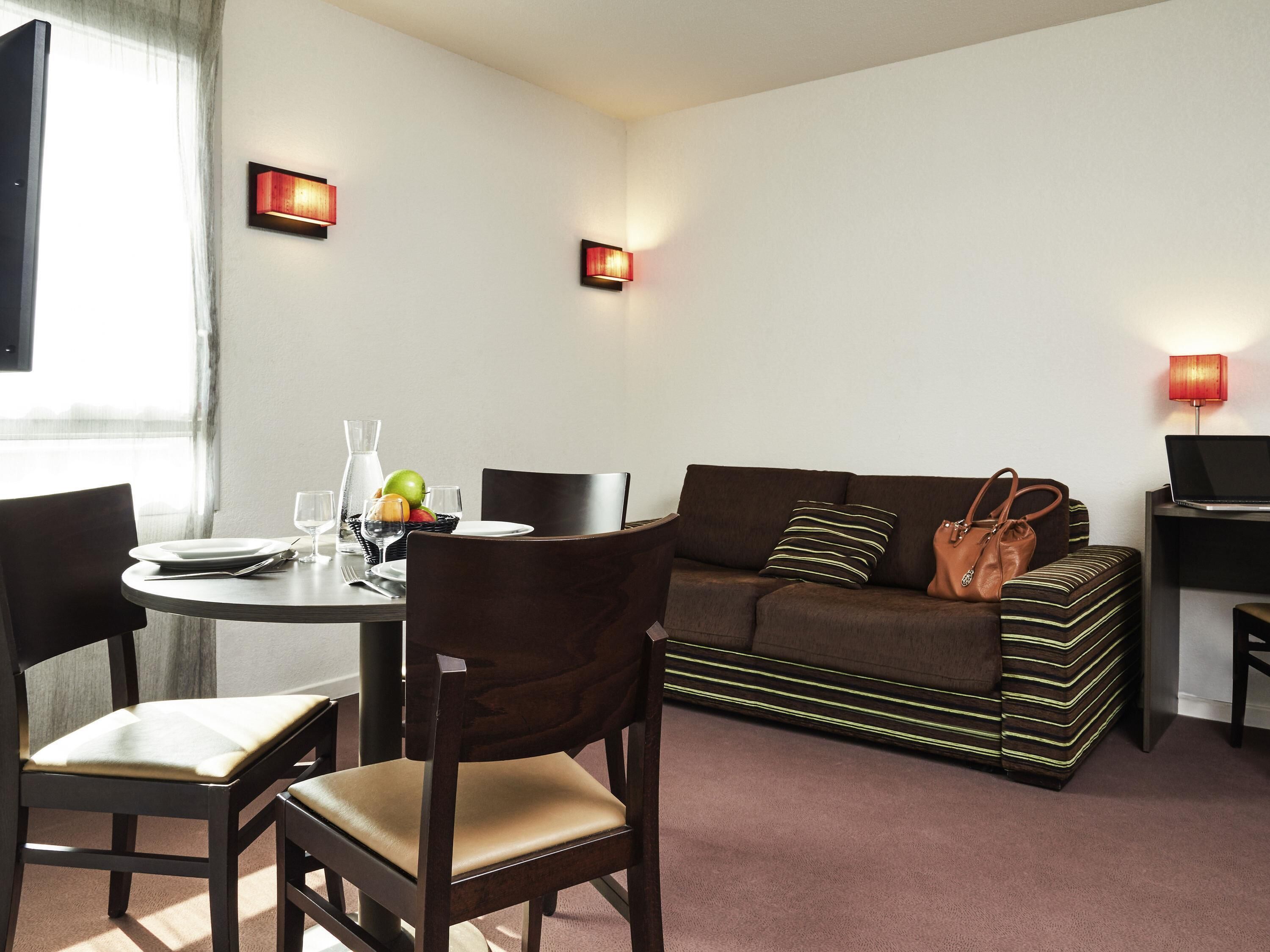 A modern living room with a dining table set for two, a sofa with a striped pillow, and a laptop on a side table.