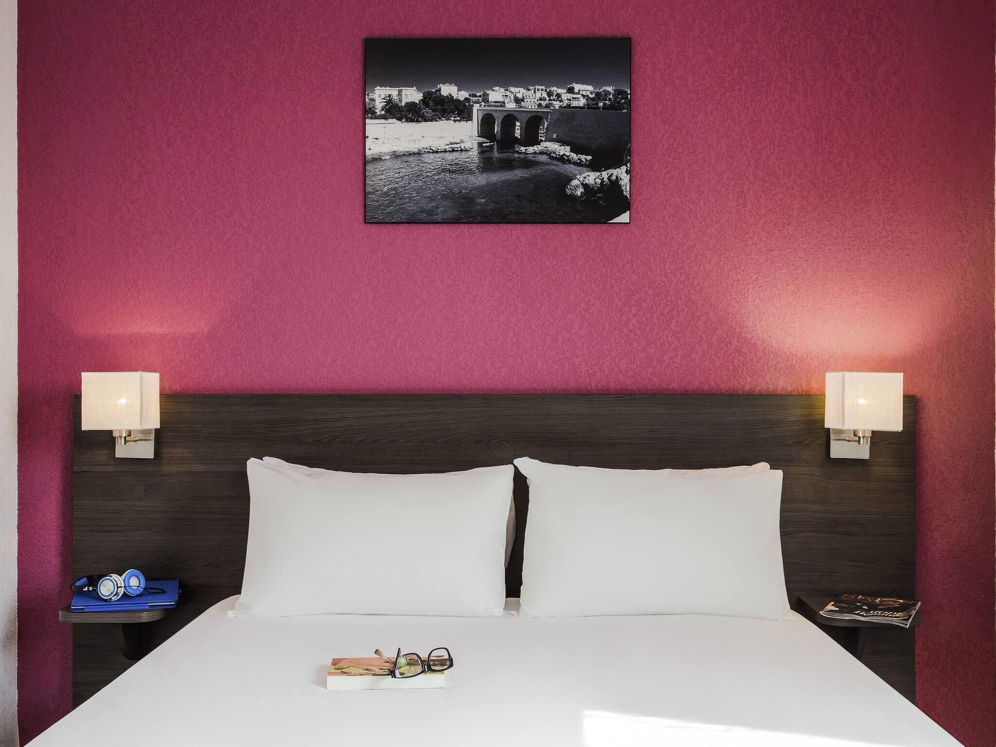 A hotel room with a bed, two bedside lamps, a nightstand with a book and glasses, and a framed picture on the wall.