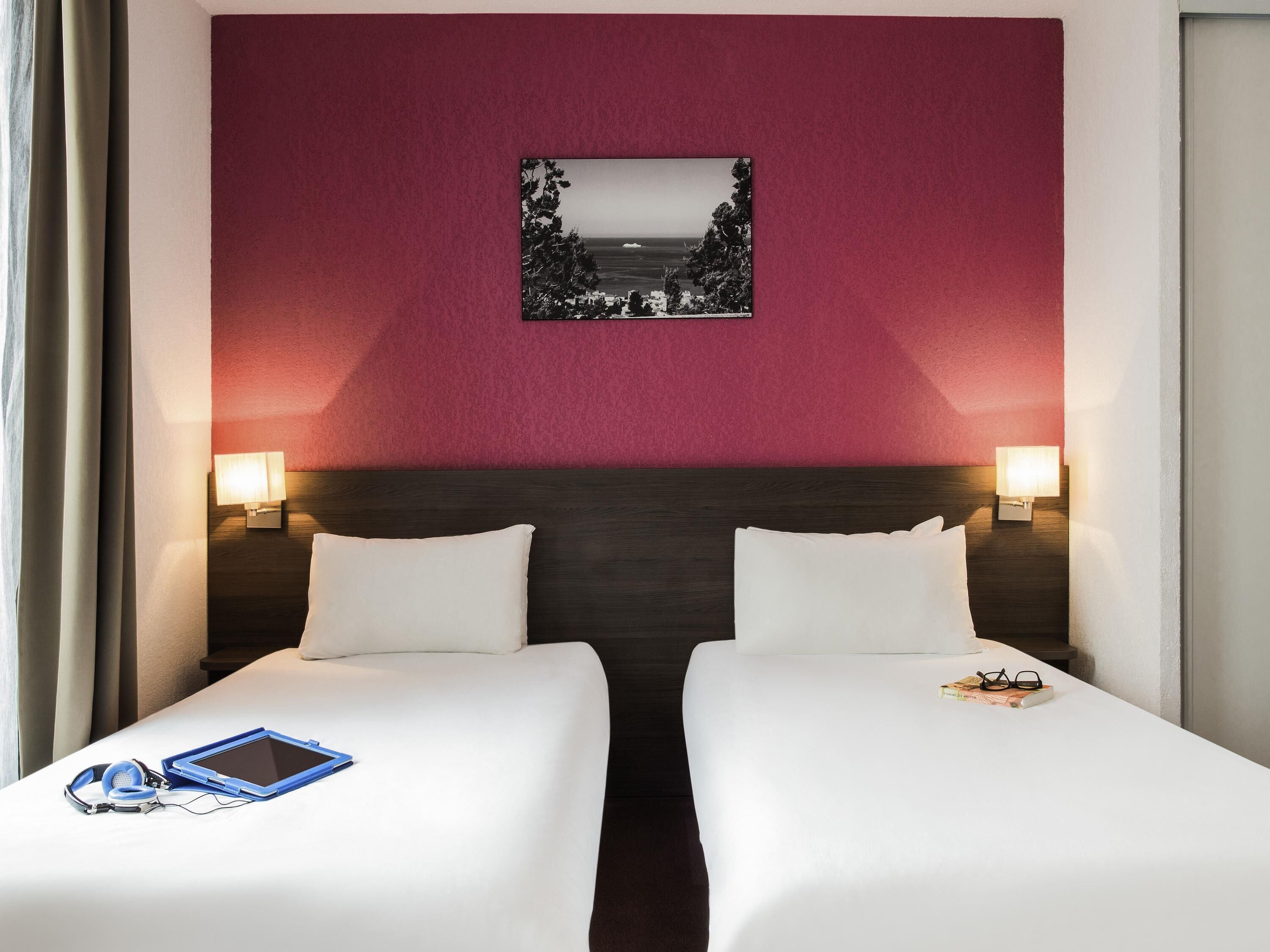 A hotel room with two beds, a nightstand with a lamp, a framed picture on the wall, and a window with curtains.