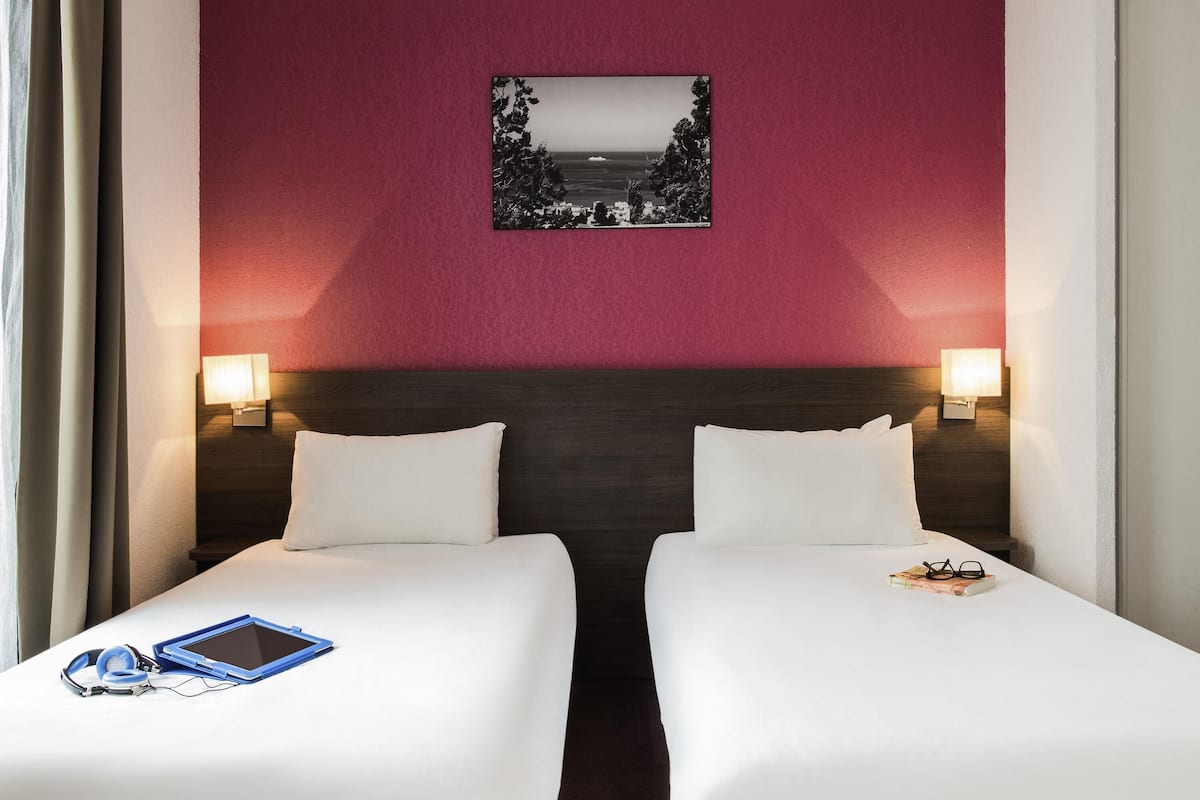 Une chambre d’hôtel avec deux lits, une table de chevet avec une lampe, un tableau encadré au mur et une fenêtre avec des rideaux.
