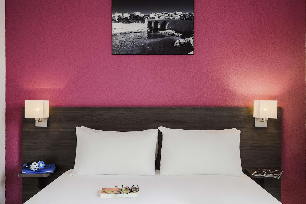 Una habitación de hotel con una cama, dos lámparas de noche, una mesita de noche con un libro y auriculares, y un cuadro en la pared.